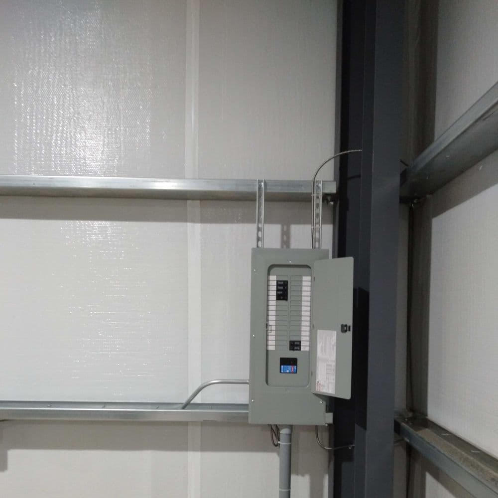Electrical panel mounted on a wall in a storage facility, with visible wiring and shelving.