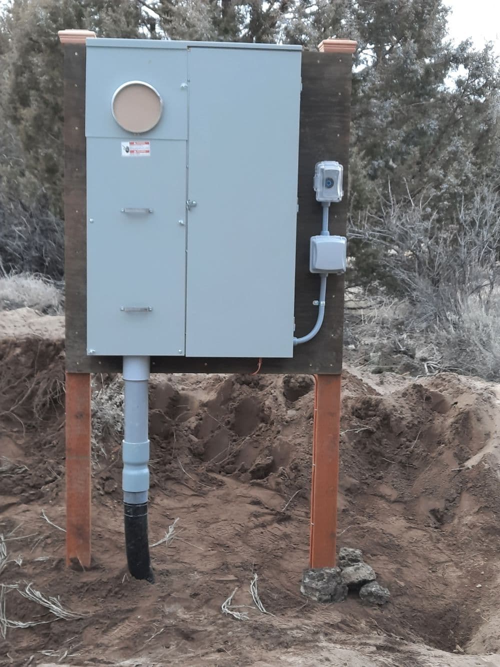 Utility control cabinet on a wooden post surrounded by sandy soil and shrubbery.