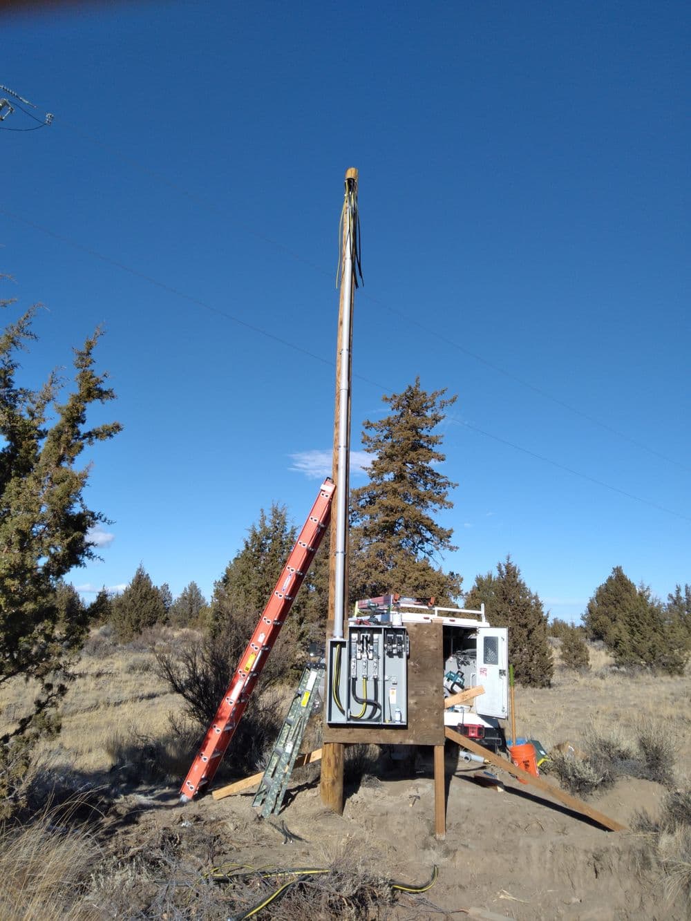 Utility pole with electrical equipment and ladder against a clear blue sky in a rural landscape.