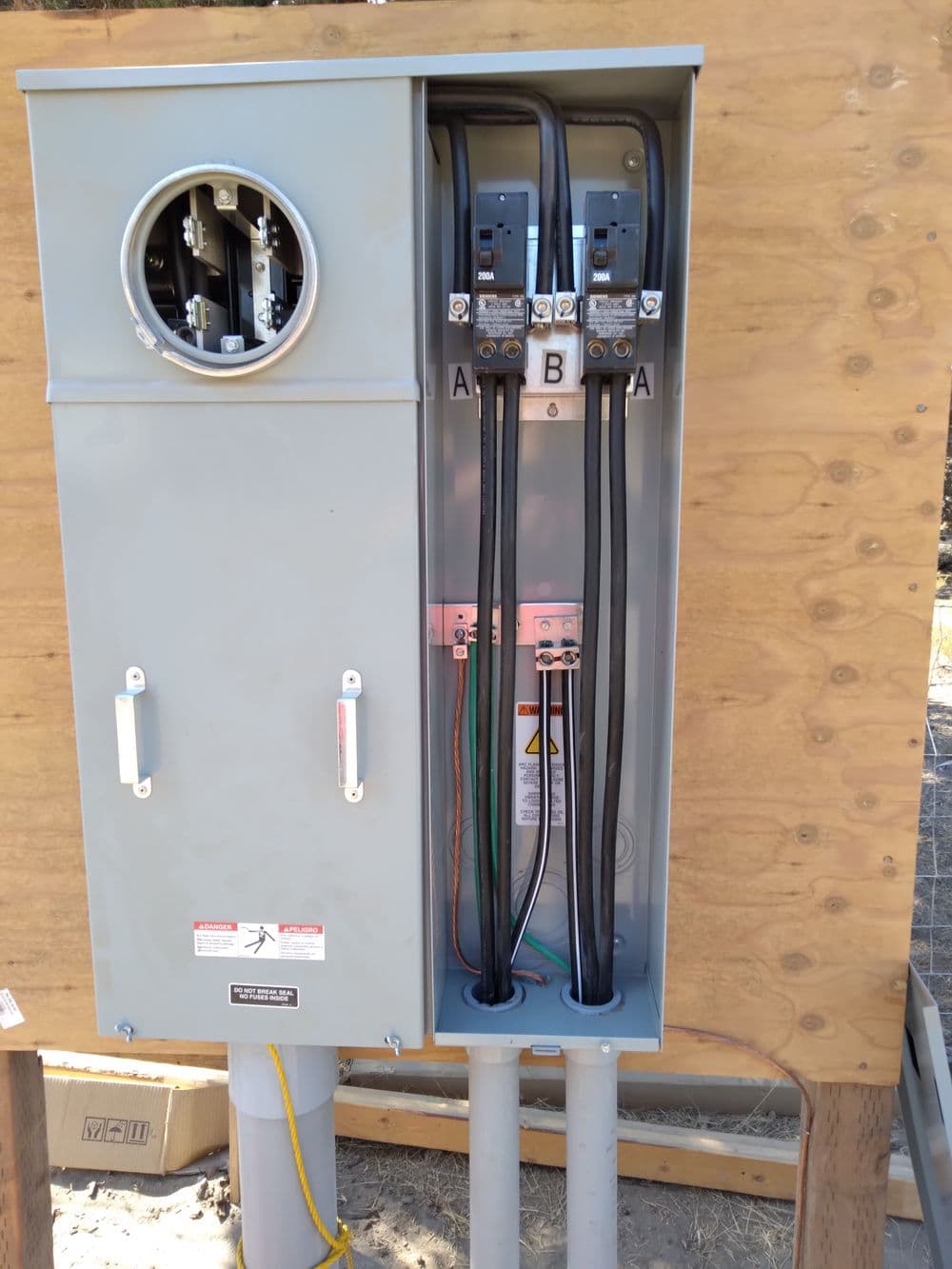 Electrical service panel with three cables, mounted on a wooden structure.