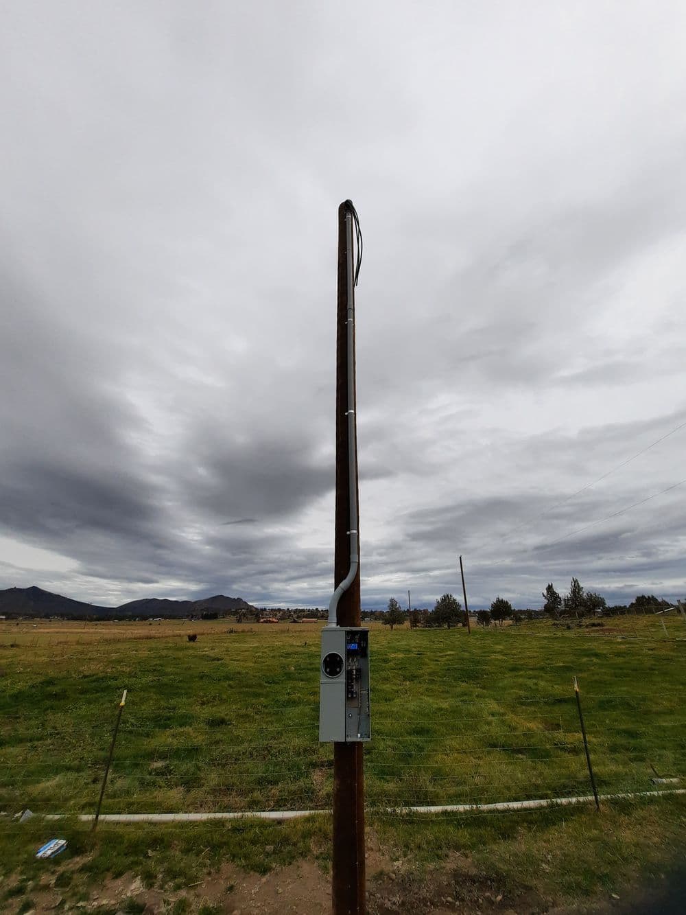 Utility pole with electrical box against a cloudy sky and grassy landscape.