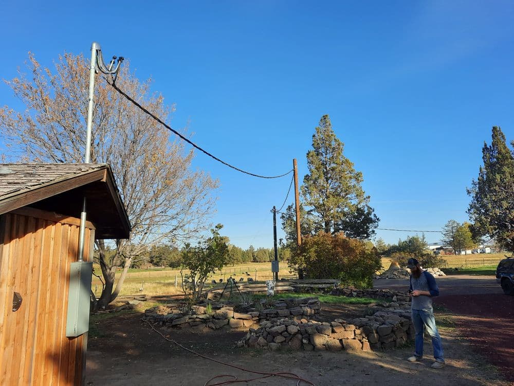 Man standing next to a wooden building with power lines and trees in a rural landscape.