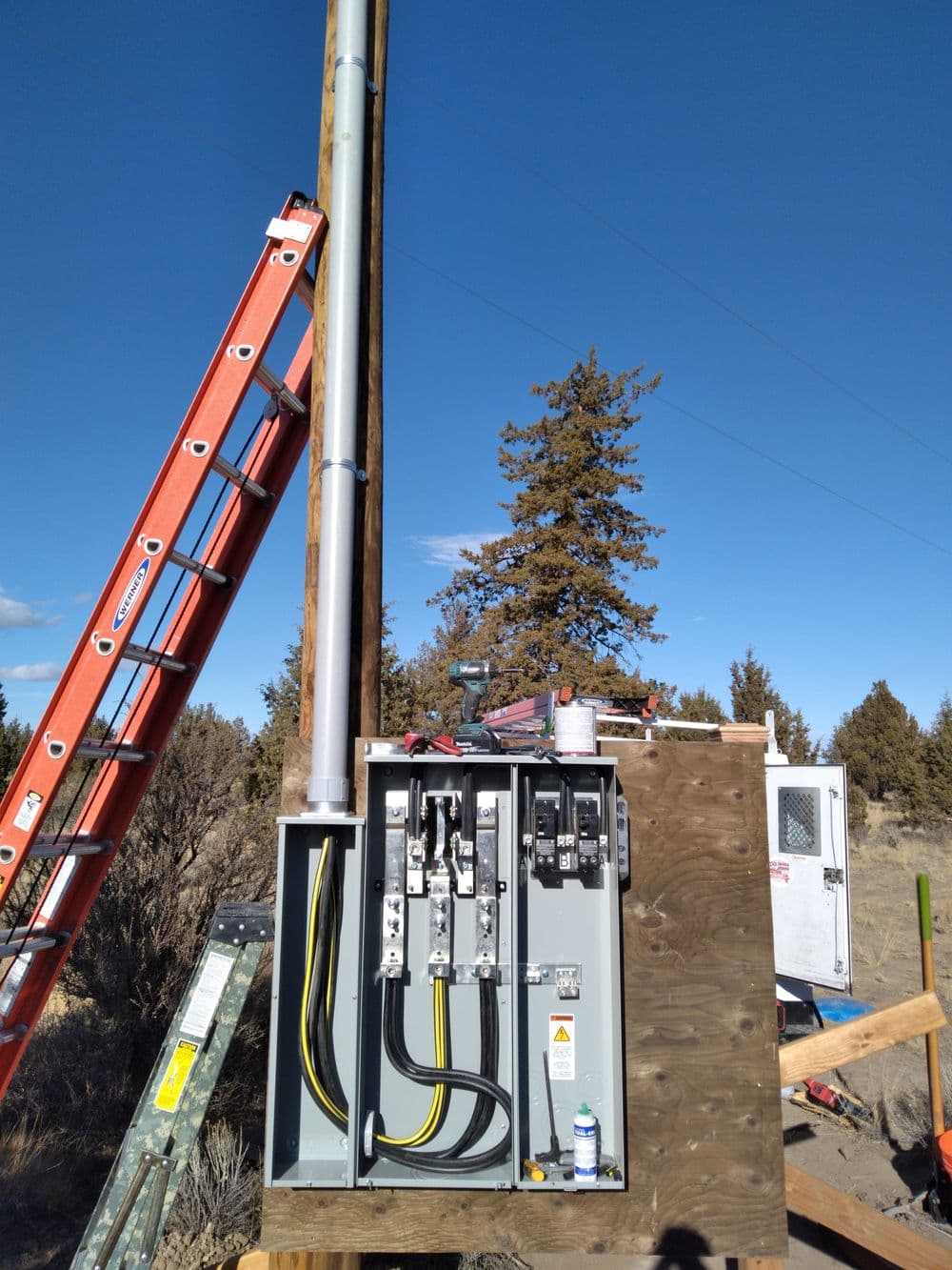 Electrical panel with power lines, ladder, and pole under a clear blue sky.