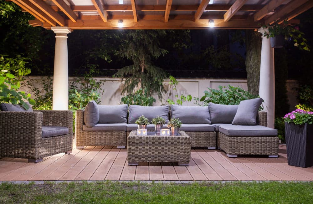 Cozy outdoor seating area with gray cushions, wooden pergola, and soft lighting at night.